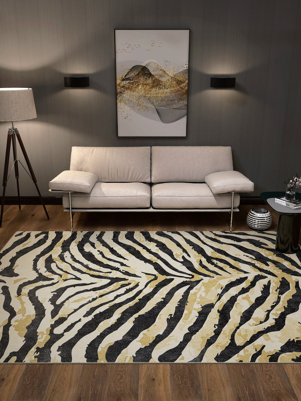 A modern living room with a beige sofa, tripod floor lamp, abstract art, and the Eternal Weave - Washable Rug - JRN39 in a black, white, and gold zebra pattern—ideal for high-traffic spaces—on wooden floors against dark walls.