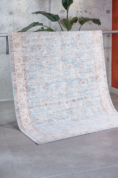 Load image 1 in gallery view. The Faded Azure - Area Rug - APX2941, featuring a subtle blue and beige pattern, is draped over a metal bar in a modern room with a concrete wall, large green plant, and gray tile floor—ideal as a stylish living room rug.