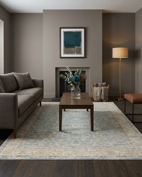 Load image 1 in gallery view. A modern living room with a gray sofa, a wooden coffee table with flowers, the Faded Azure - Area Rug - APX2941, a floor lamp, a basket of blankets, and abstract art above the fireplace.