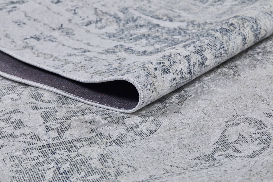 A close-up of the Faint Memories - Washable Rug - JR1437 (Custom Size), folded to display its light gray flat weave, subtle distressed dark gray pattern on a light background, highlighting texture and weave detail.