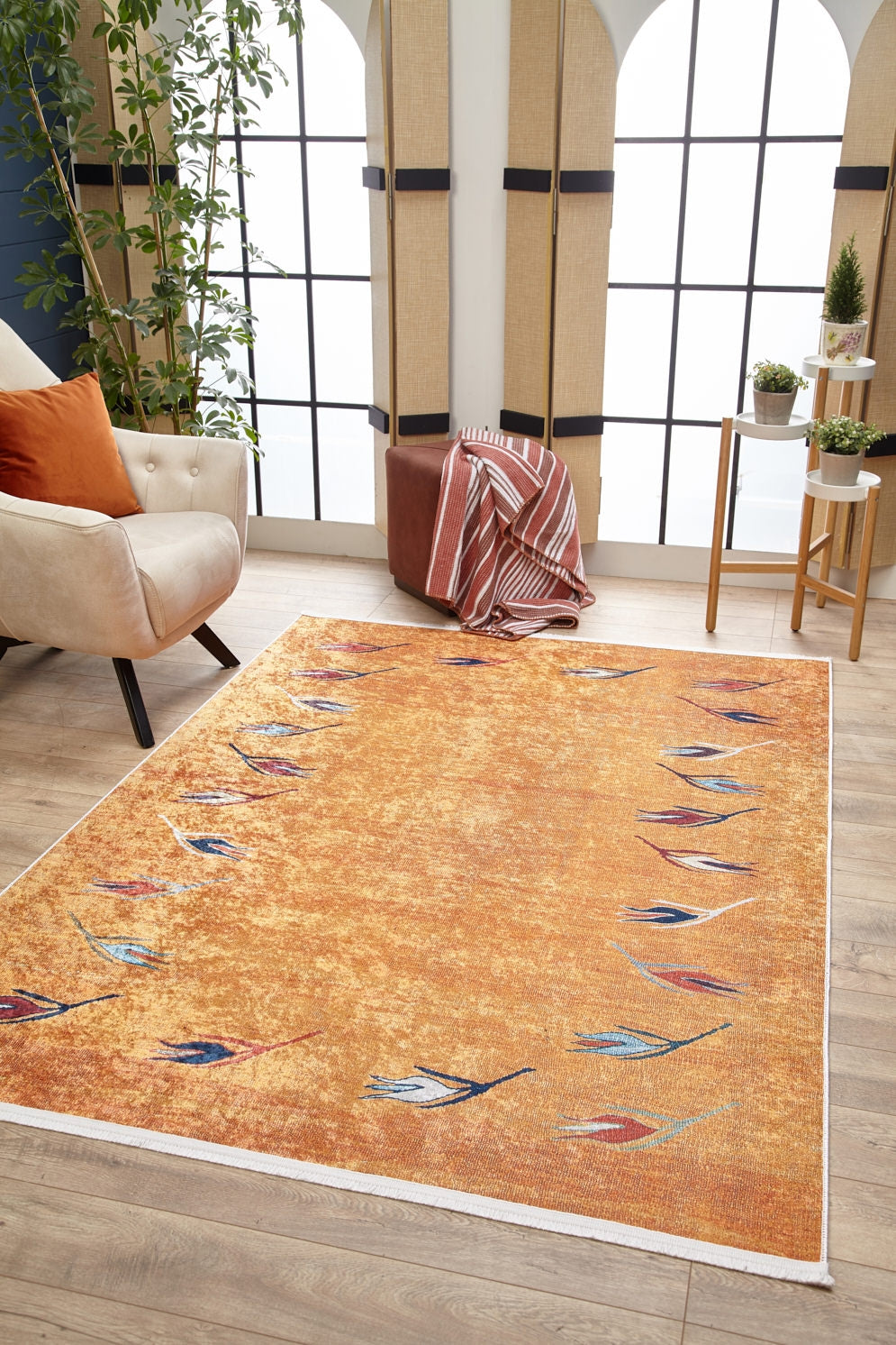 A cozy living room features the Floral Mosaic - Washable Rug - JR1720 (Custom Size) in orange with leaf patterns, a beige armchair with an orange pillow, striped blanket, brown pouf, potted plants, and large arched windows for natural light.