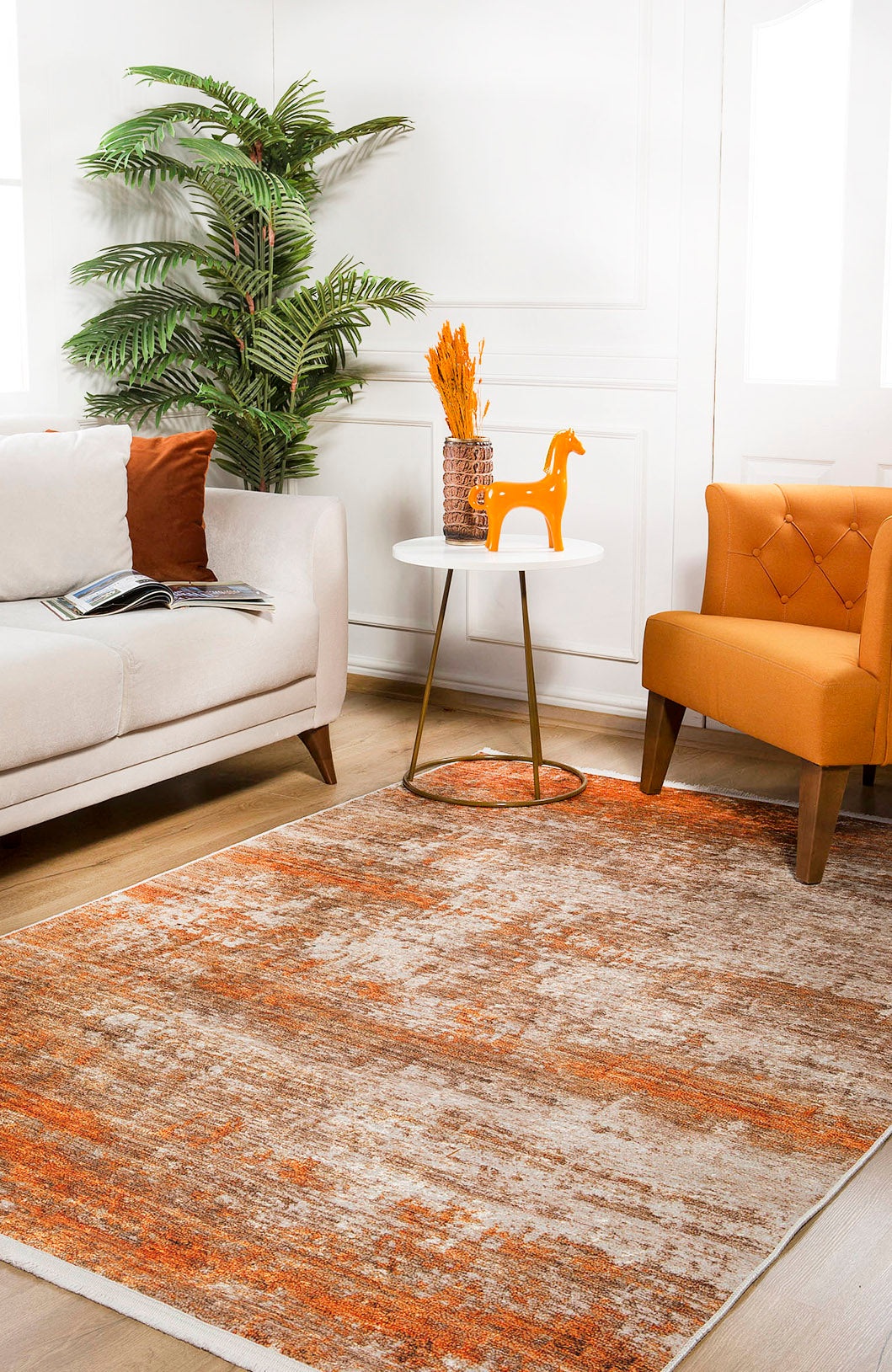 A cozy living room features a beige sofa, orange armchair, round side table with decor, potted plant, and the Fluffy Brushed - Washable Rug - JR584 (Custom Size) in orange on light wood flooring—ideal for high-traffic areas. Sunlit windows brighten the space.