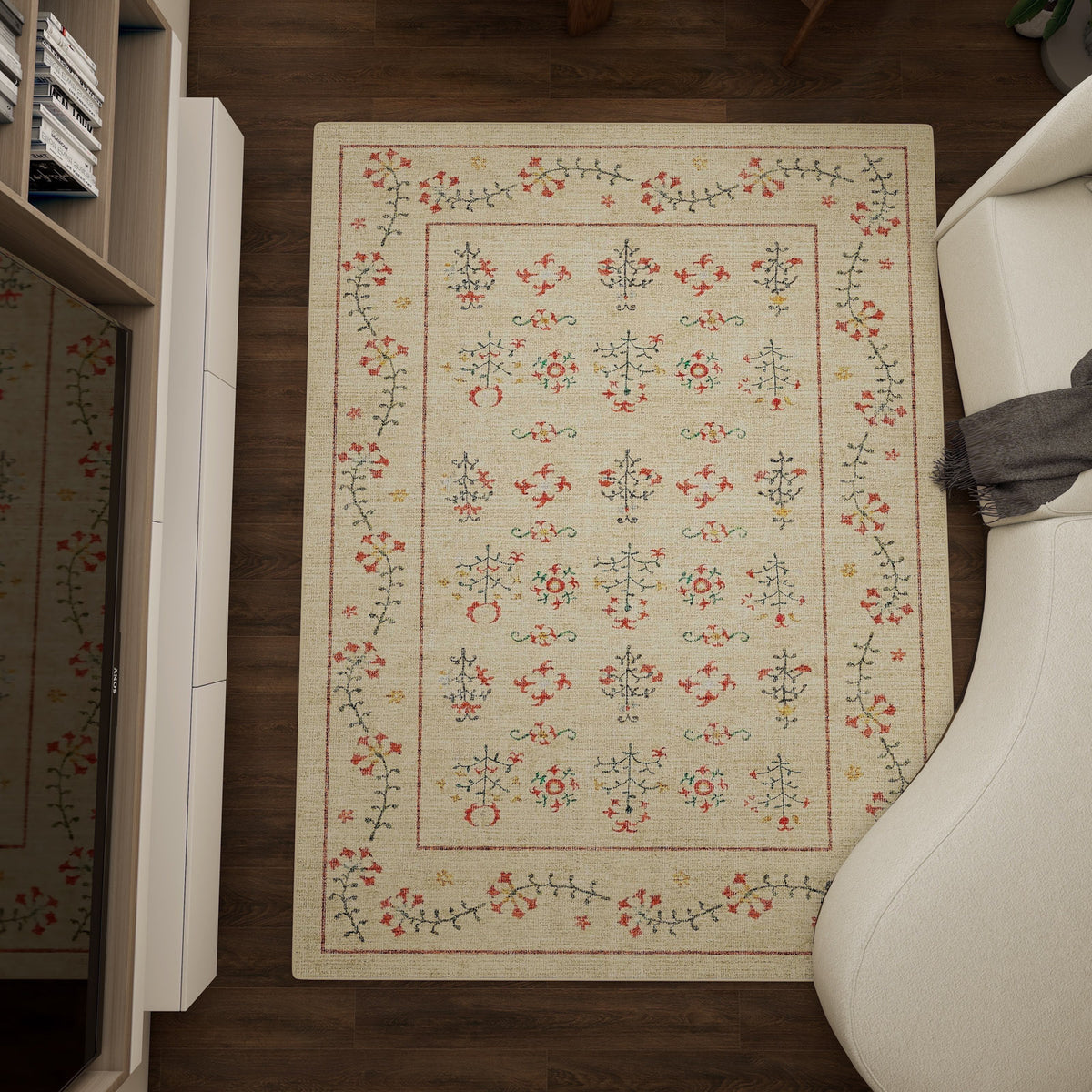 The Garden Cream - Washable Rug (JRN1026), a rectangular beige rug with red, green, and black floral patterns, is placed on a dark wooden floor beside a white curved sofa and a light-colored cabinet.