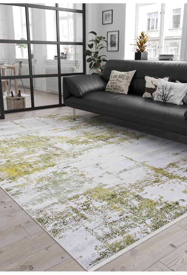 A modern living room features a black sofa, decorative pillows, plants, and large windows. The Geometric Geometry - Washable Rug - JR1290 in green adds a pop of color, while neutral tones and minimal decor create a cozy, stylish vibe.