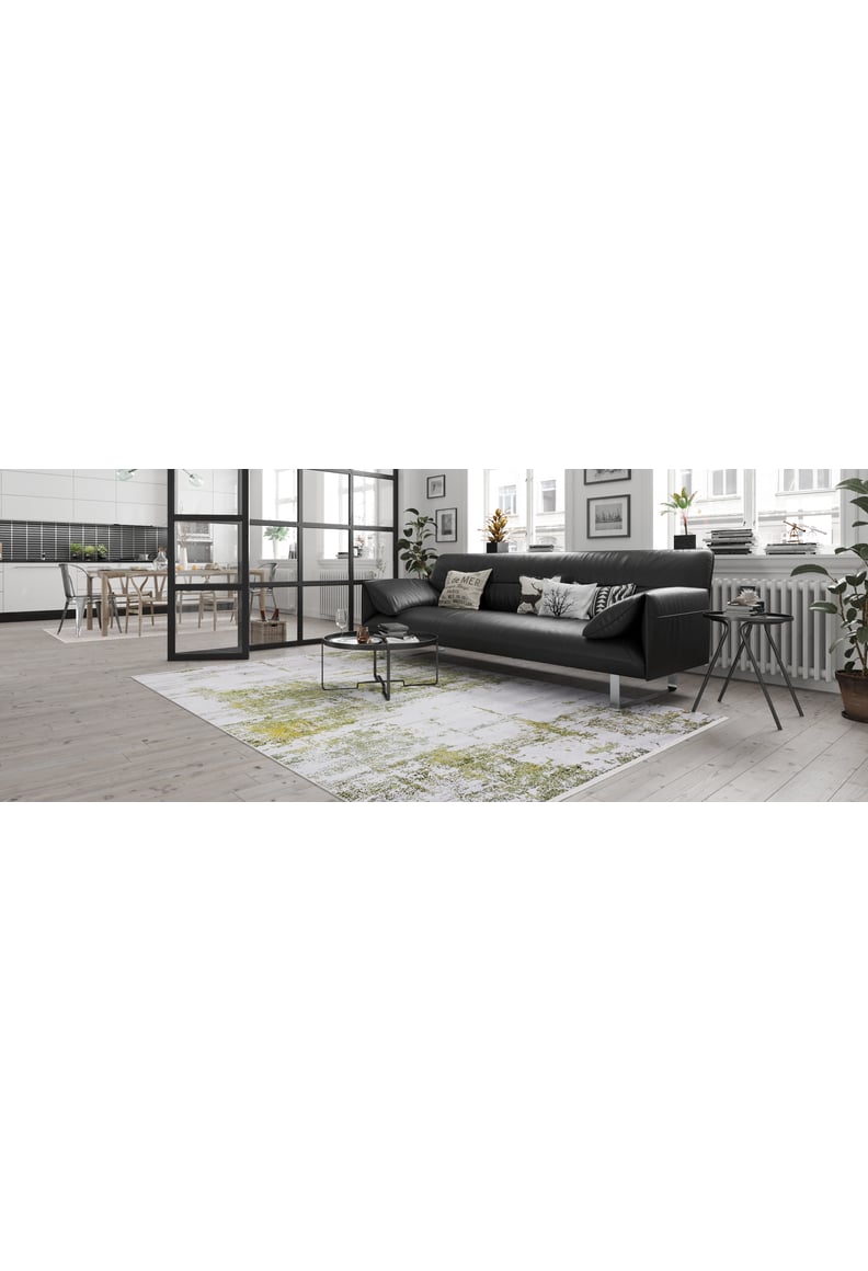 Modern living room with a black sofa, the Geometric Geometry - Washable Rug - JR1290, and a small round coffee table. Large windows and a glass wall divide the space. Light wood flooring and indoor plants create warmth.