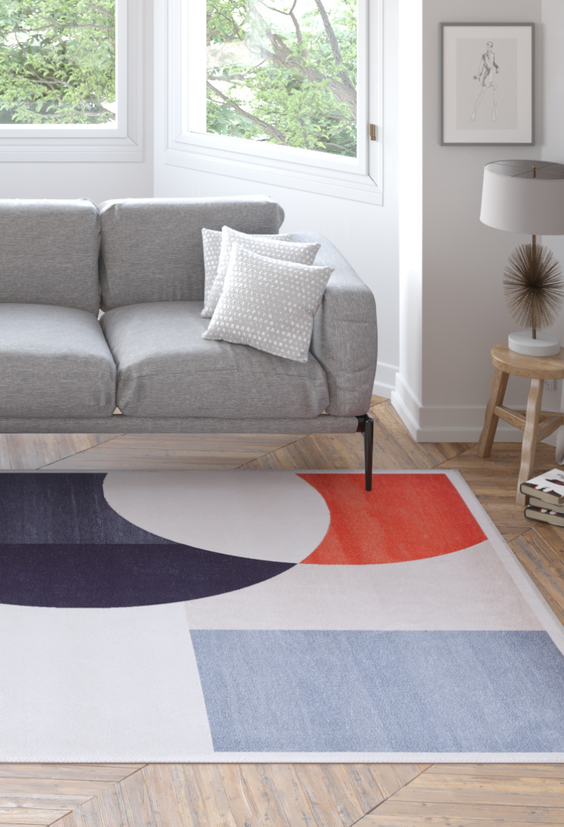 A modern living room featuring a light gray sofa, a white pillow with gray dots, large windows with greenery outside, a side table with a lamp and framed art, and the Geometric Zen Modern Washable Rug - MDJ002 in blue, black, red, and white patterns.