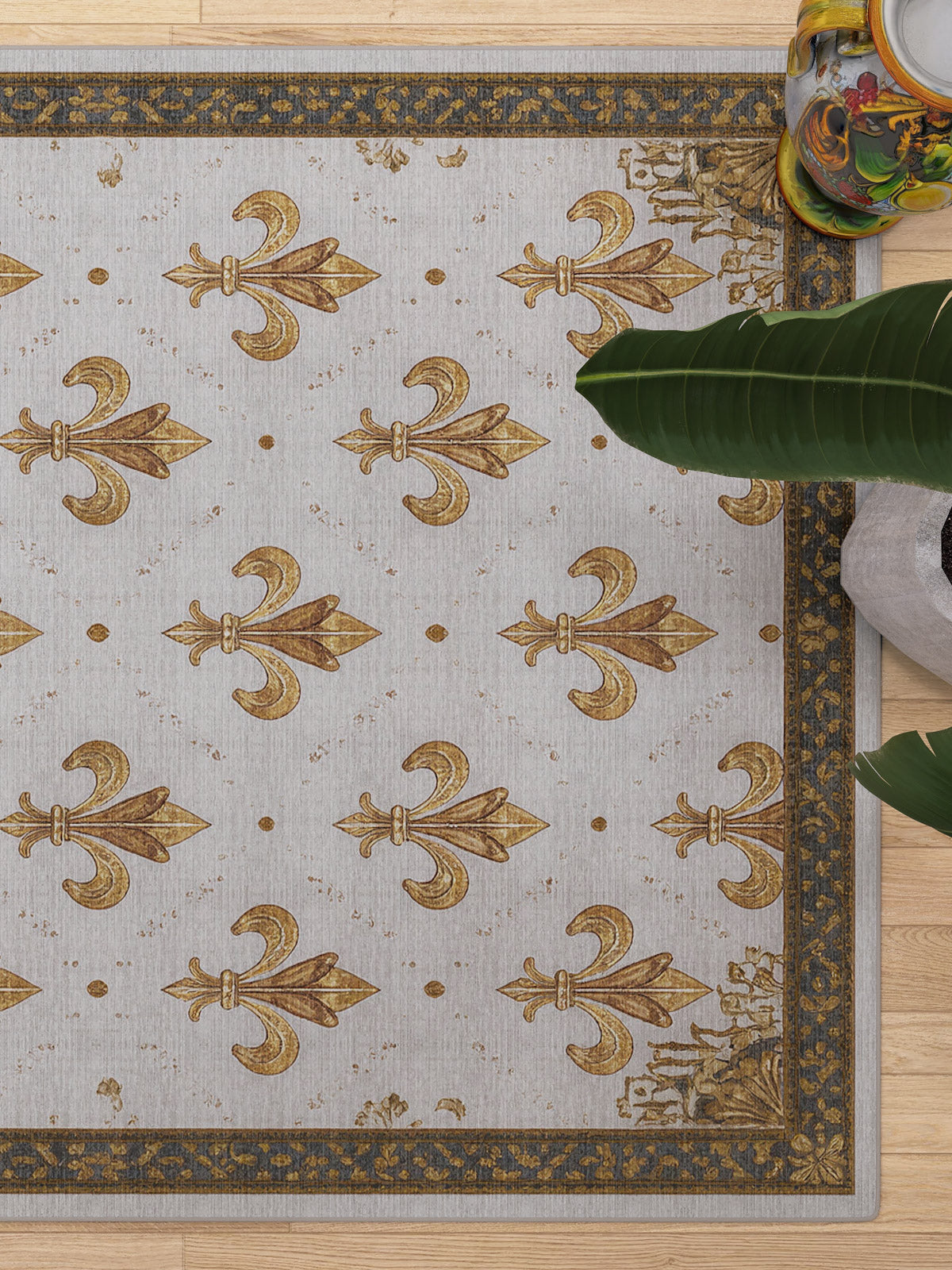The Golden Crest - Washable Rug - JRS015 (Custom Size), featuring a gold fleur-de-lis design and border, lies on a wooden floor—perfect for allergy-sensitive homes. Green leaves and colorful pottery accent the top right corner.