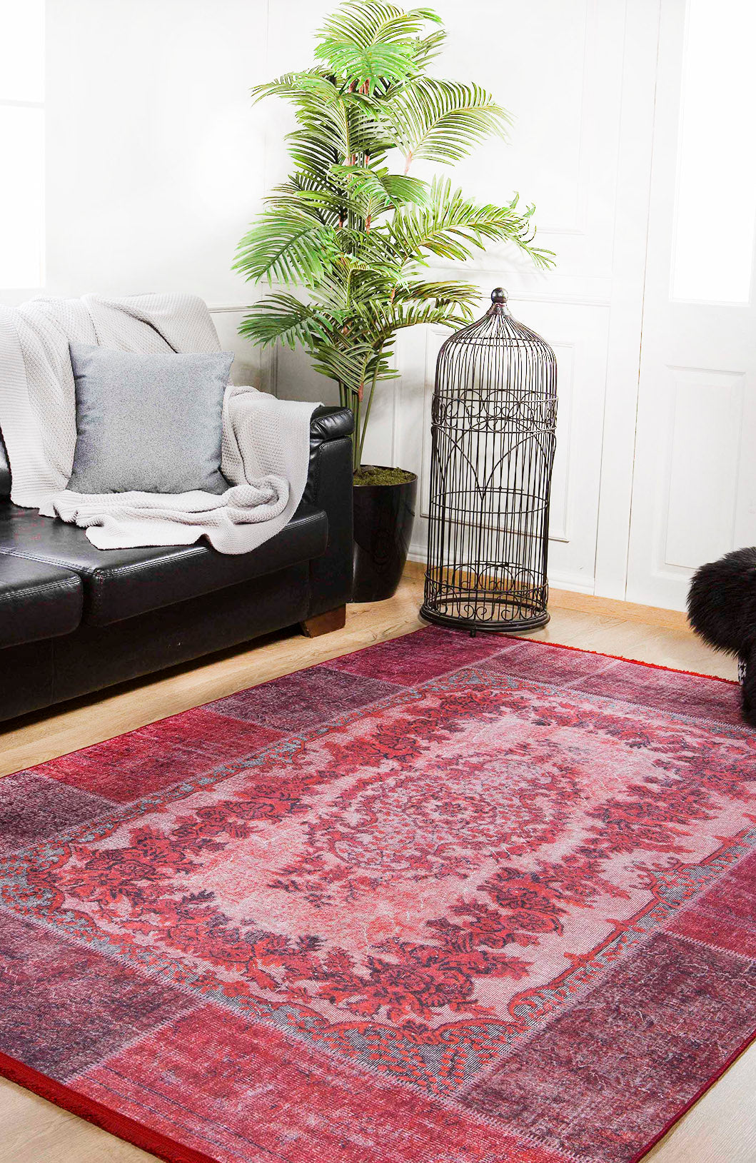A cozy living room with a black couch, gray blanket and pillow, the Golden Wonder - Washable Rug - JR306 (Custom Size) in red—ideal for high-traffic areas—a tall potted plant, decorative birdcage, and white walls with natural light.
