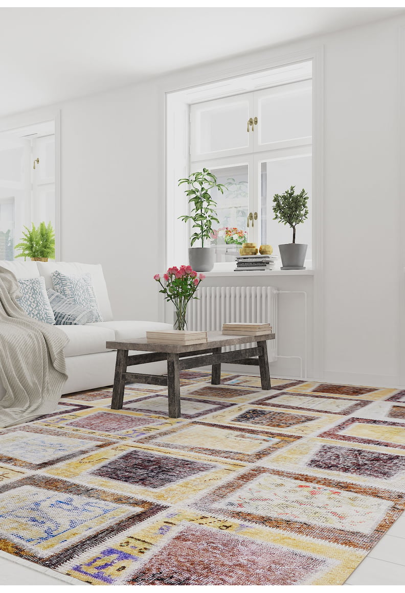 A bright living room features a white sofa with blue and white pillows, a wooden coffee table with books and pink flowers, plants on the windowsill, and the Gridiron Glow - Washable Rug - JR183 (Custom Size) ideal for high-traffic areas.