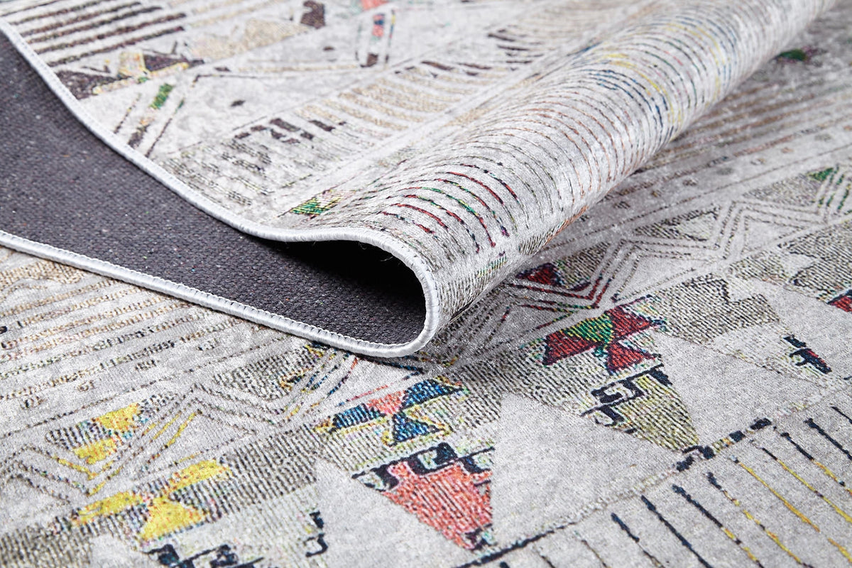A close-up of the Gypsy Glam - Washable Rug - JR1716 (Custom Size) shows its geometric patterns and colorful accents on a light backdrop, with one corner folded to reveal the textured underside.