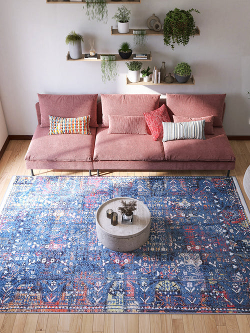 Load image 1 in gallery view. A modern living room with a pink sectional sofa, colorful pillows, and a round concrete coffee table. The space is anchored by the Gypsy Glitz - Washable Rug - JR1650, ideal for high-traffic areas. Shelves of green plants are mounted above the sofa.