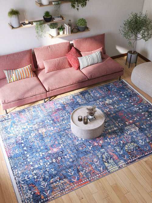 Load image 1 in gallery view. A bright living room features a pink sectional sofa, colorful cushions, the Gypsy Glitz - Washable Rug - JR1650 in blue for high-traffic areas, a round coffee table, wooden floors, potted plants, and wall-mounted shelves with greenery.