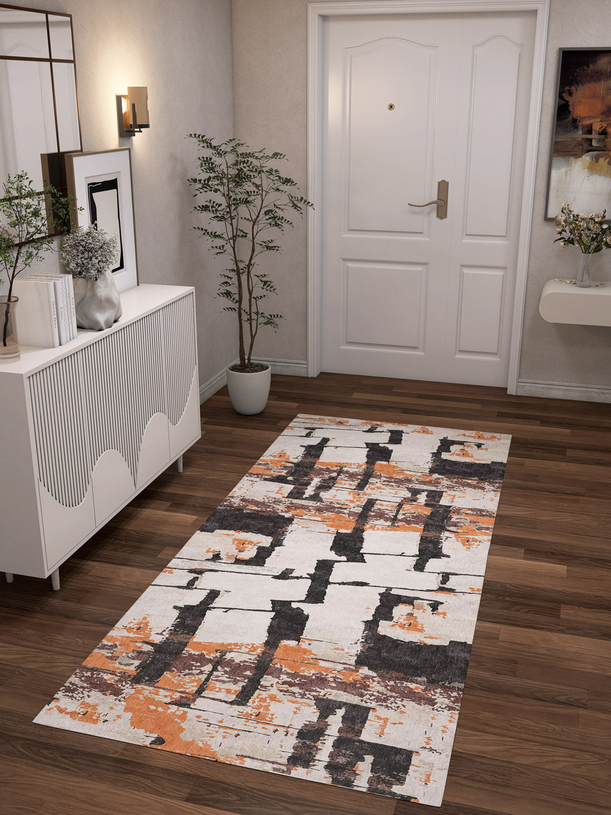 A modern hallway with a white front door, wood floors, and the Heirloom Touch - Washable Rug - JRN42 (Custom Size) in white, black, and orange. A white console, plant, and art on beige walls complete this chic space—ideal for high-traffic areas.