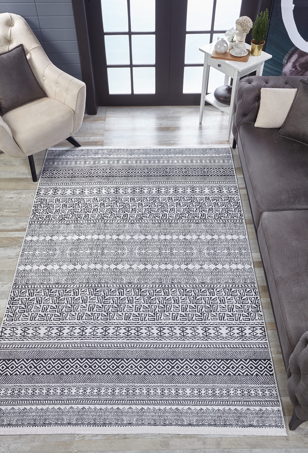 A living room featuring the Indie Impressions - Washable Rug - JR1648 (Custom Size) in gray-anthracite, a beige armchair, gray sofa, white side table with plants and decor, and large glass doors allowing natural light in.