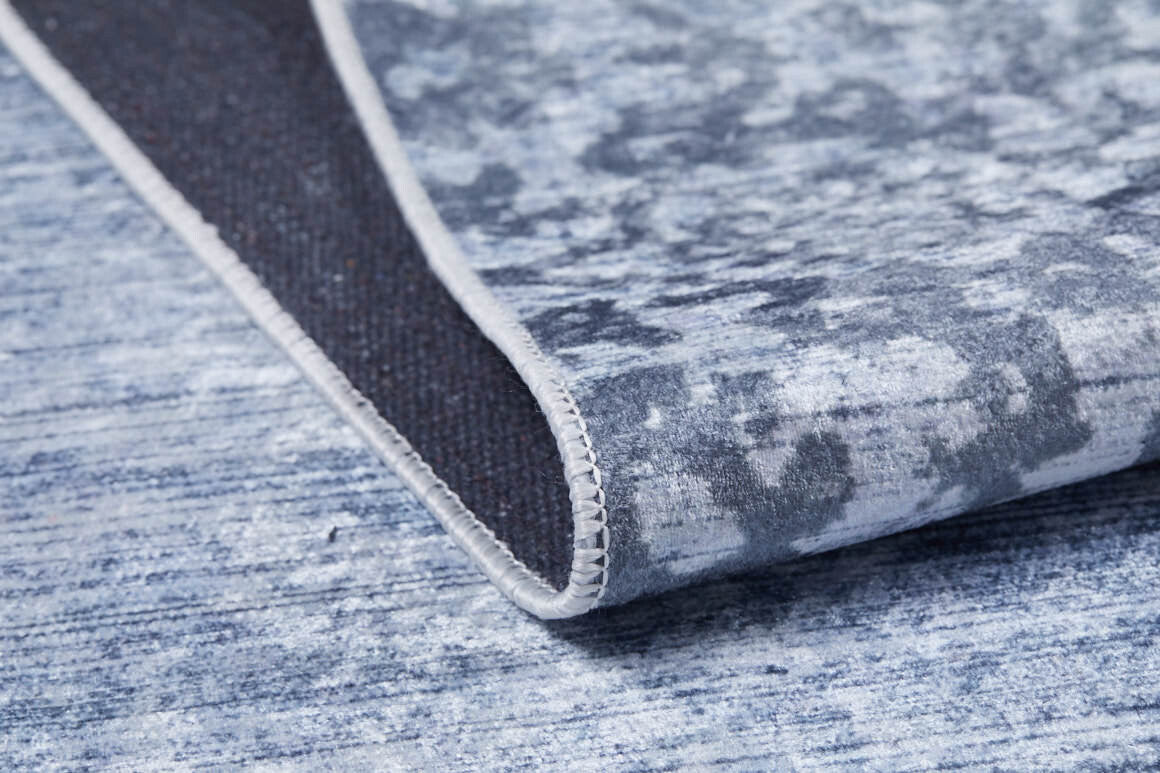 A close-up of the Intricate Scroll - Washable Rug - JR342 (Custom Size) in blue and gray, partially lifted to display its dark underside and fine edge stitching—perfect for allergy-sensitive spaces or busy areas.