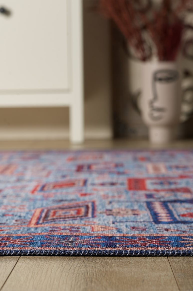 A close-up of the Birdwatcher's Dream Blue Washable Rug - LCC3020 (Custom Size) on a wooden floor, beside a white cabinet and decorative face vase—ideal for high-traffic areas.
