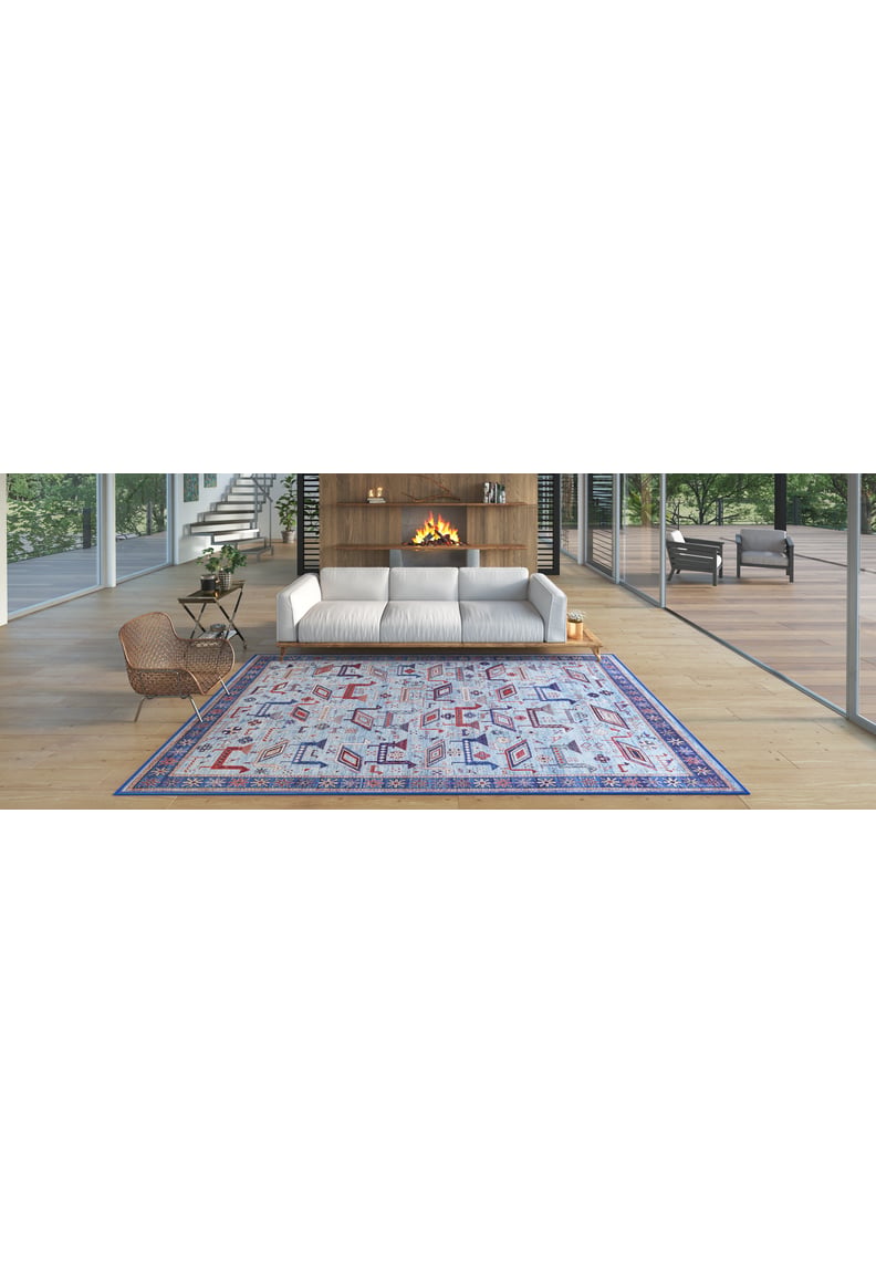 A modern living room features a white sofa, round chair, fireplace in a wooden wall, and the Birdwatcher's Dream Blue Washable Rug - LCC3020 (Custom Size). Large glass windows overlook trees and a spacious deck—perfect for high-traffic areas.
