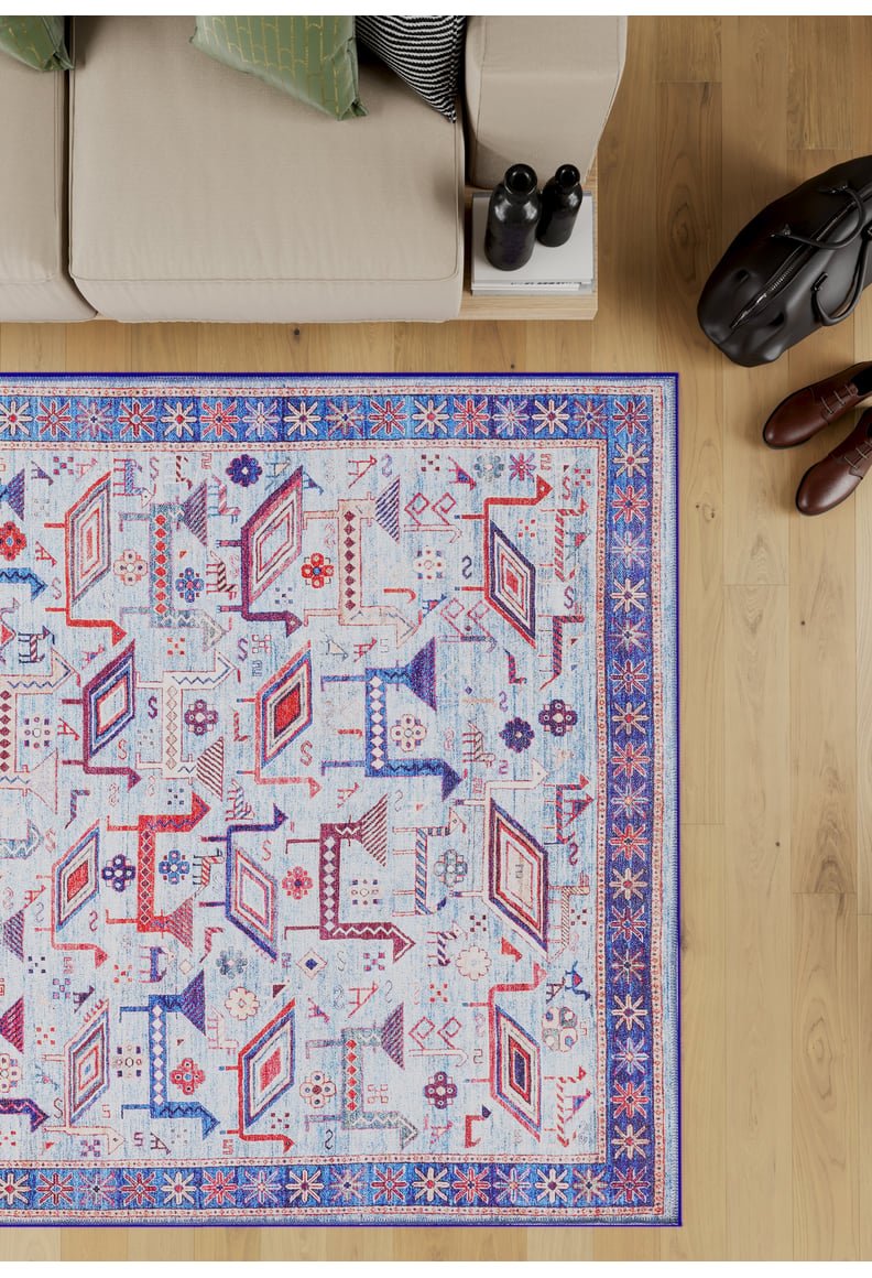 The Birdwatcher's Dream Blue Washable Rug - LCC3020 (Custom Size), with geometric blue, red, and orange patterns and a decorative border, lies on a light wood floor beside a beige sofa, black bag, and brown shoes—ideal for high-traffic areas.