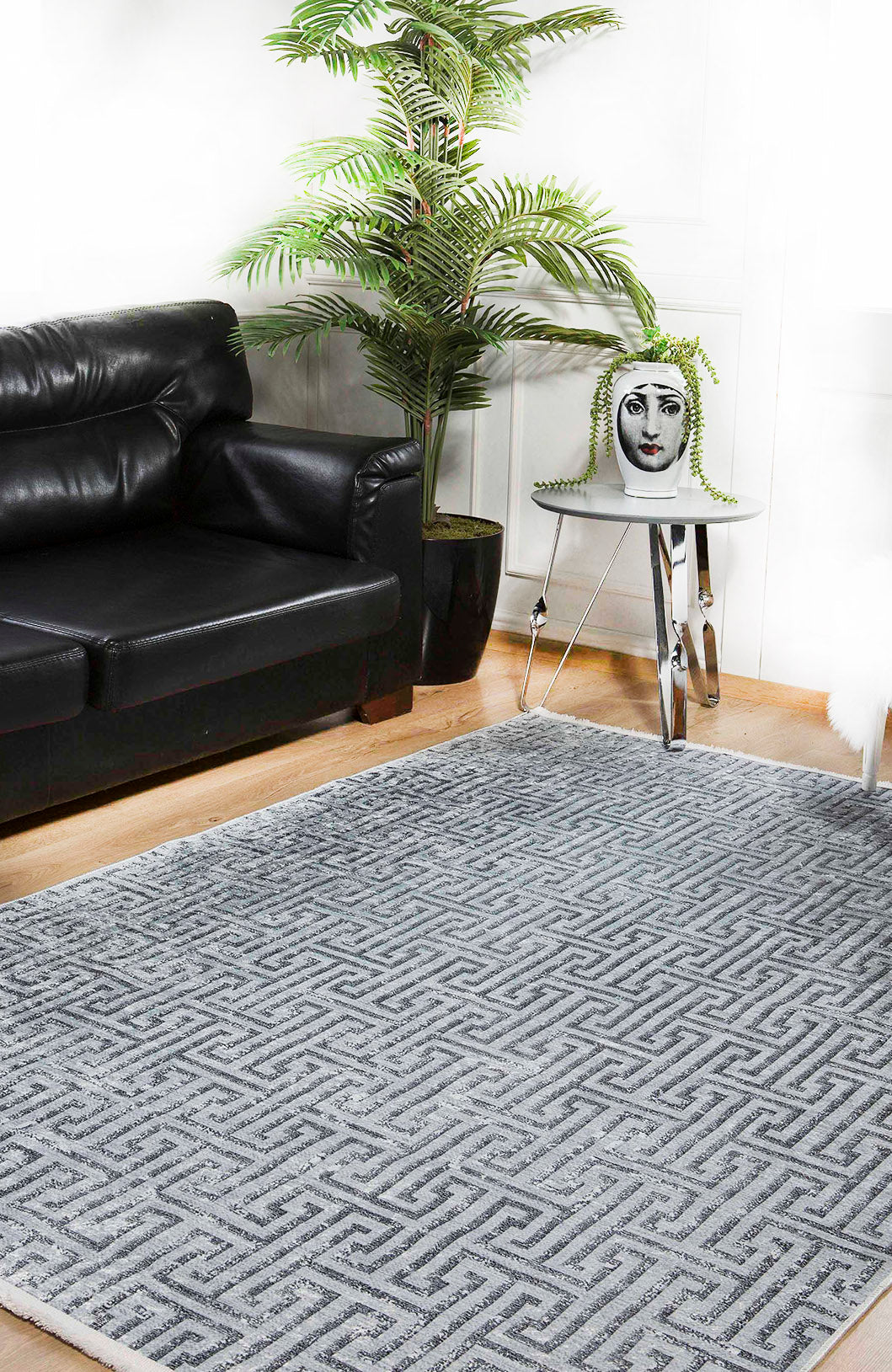 A modern living room features a black leather sofa, green potted plant, small metal side table with a decorative vase of a woman's face, and the Jazz Age - Washable Rug - JR1225 (Custom Size) in gray-anthracite on the wooden floor.