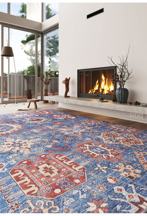 Load image 1 in gallery view. A cozy living room with a lit fireplace, modern decor, floor-to-ceiling windows, and the Kaleidos Authentic Blue Washable Rug—ideal for high-traffic spaces. Sculptures and a vase with branches sit on the mantel.