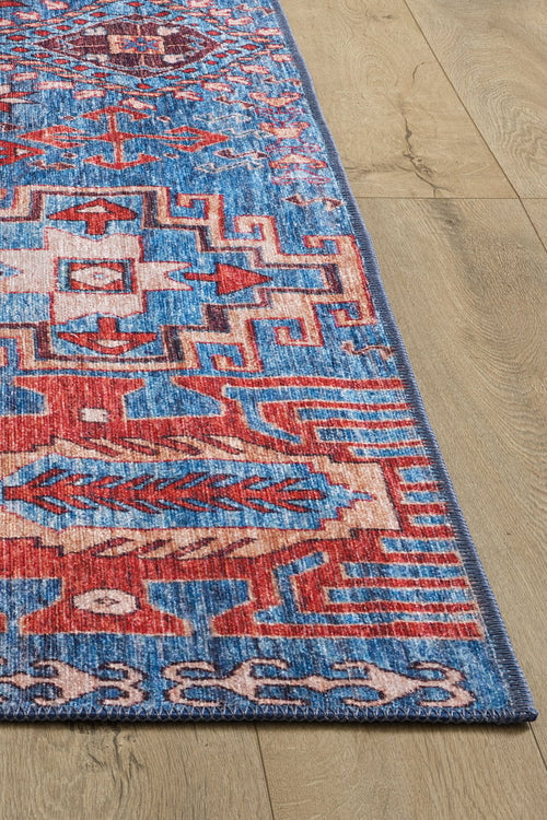 Load image 1 in gallery view. A close-up of the Kaleidos Authentic Blue Washable Rug - LCC3028, featuring geometric patterns in red, white, and blue on a light wood floor. Only a corner and edge of this durable, high-traffic area rug are shown.