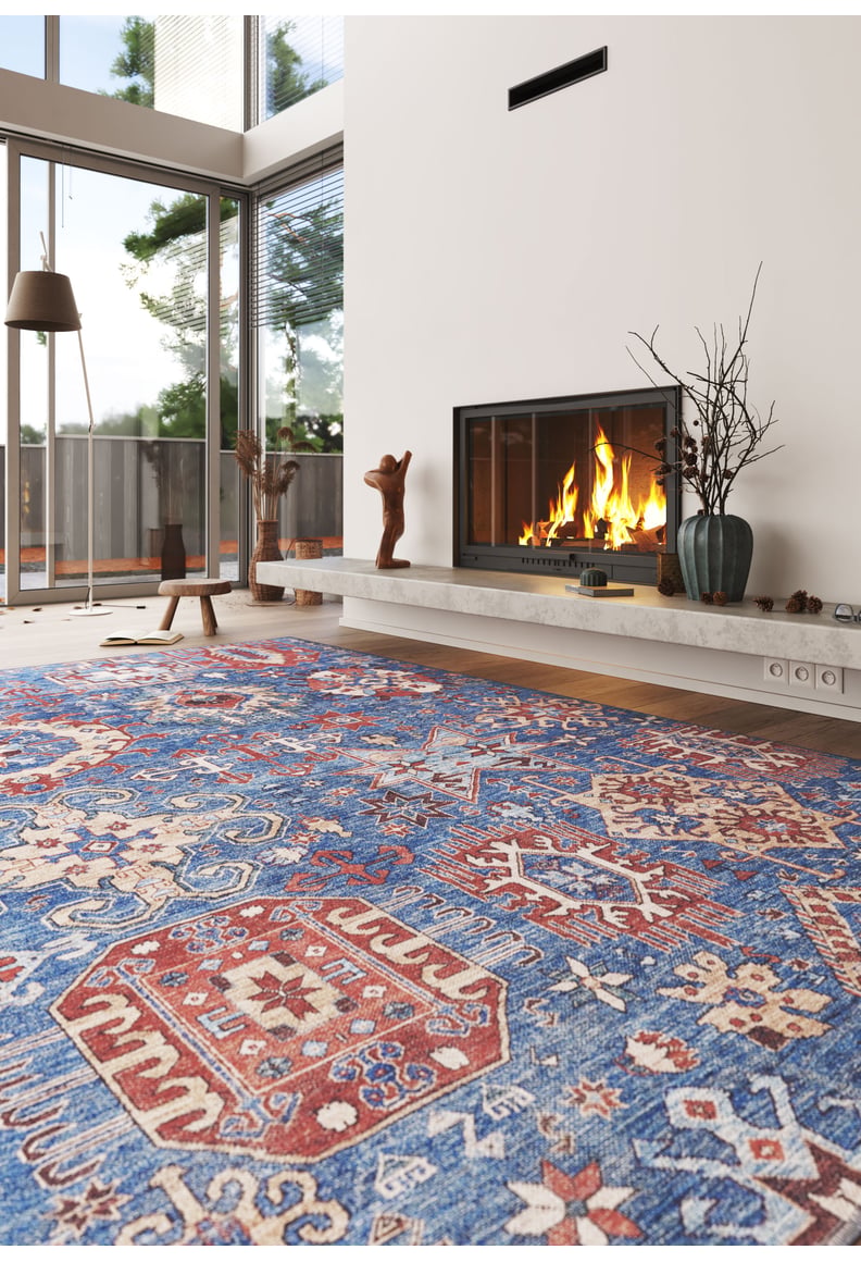 A modern living room features tall windows, a lit fireplace, minimalist décor, and a large Kaleidos Authentic Blue Washable Rug - LCC3028 (Custom Size) with colorful patterns—ideal for high-traffic areas. Decorative vases accent the mantel.