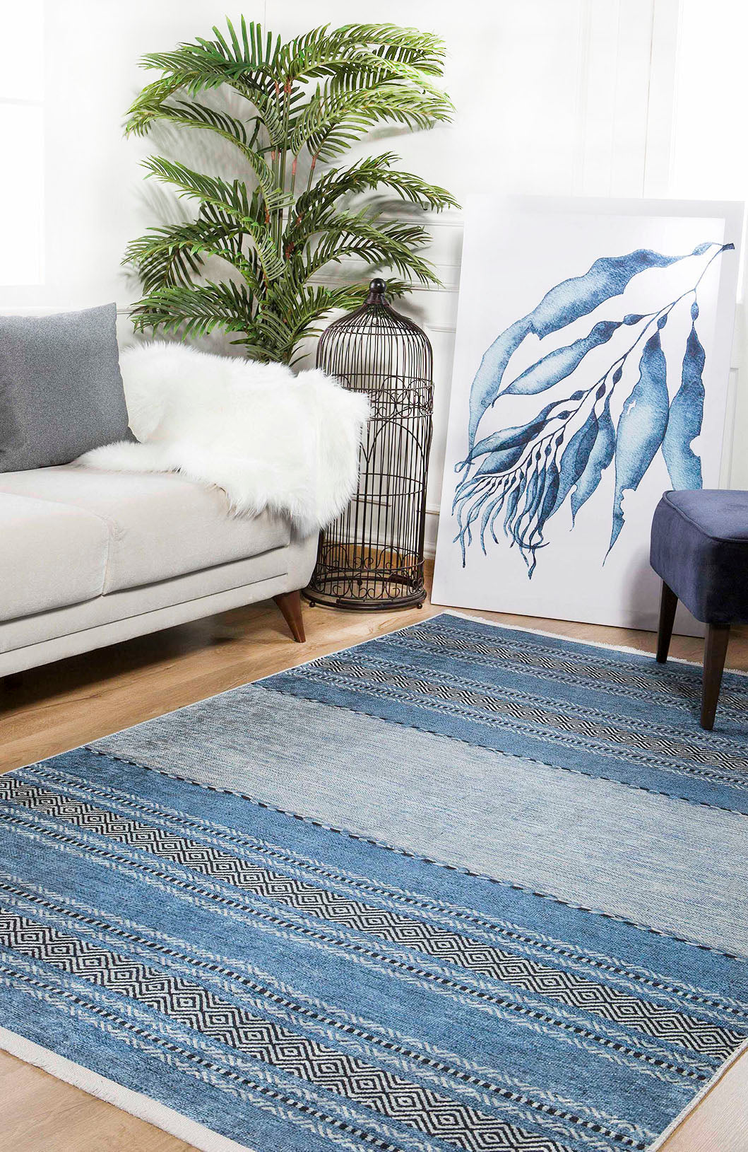A cozy living room featuring a light gray sofa, the Layered Lines - Washable Rug - JR1545 (Custom Size) in blue for high-traffic areas, a navy ottoman, a leafy potted plant, large blue abstract artwork, and a decorative birdcage.