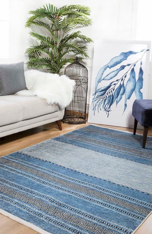 Load image 1 in gallery view. A cozy living room featuring a light gray sofa, the Layered Lines - Washable Rug - JR1545 (Custom Size) in blue for high-traffic areas, a navy ottoman, a leafy potted plant, large blue abstract artwork, and a decorative birdcage.