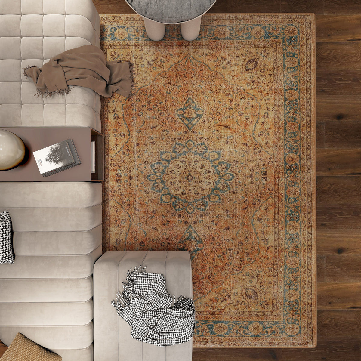 A top-down view of a living room featuring the Classic Laurel - Washable Rug - JRN1022 (Custom Size) in ornate orange and blue, with a beige sectional, small round table, books, globe on a side table, and a checkered blanket draped over the sofa.