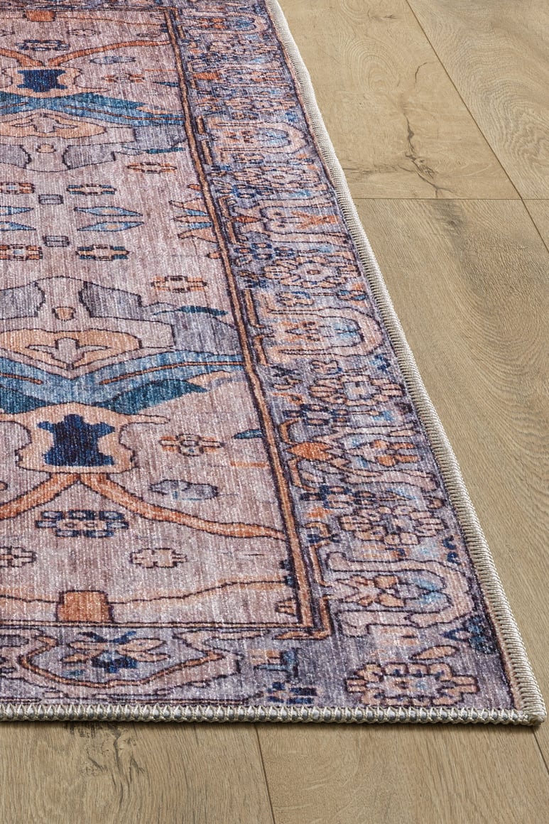 A close-up of the Luminary Beige Gray Washable Rug (LCC3023), featuring a detailed blue, beige, and pink pattern on a light wood floor, highlights the rug’s woven edge and corner.