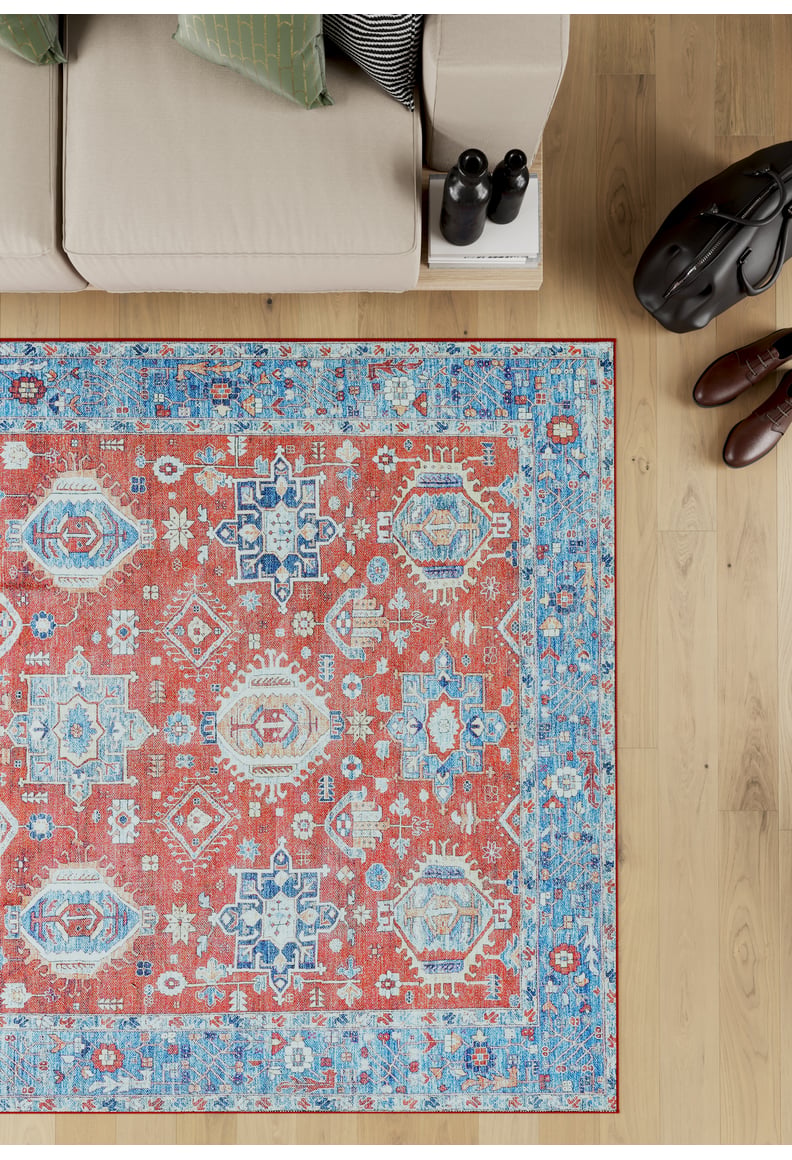 The Lunavale Red Blue Washable Rug - LCC3007 (Custom Size), featuring red, blue, and cream geometric patterns, sits on a light wooden floor beside a beige sofa—ideal for high-traffic areas. Shoes, a bag, and water bottles rest nearby.