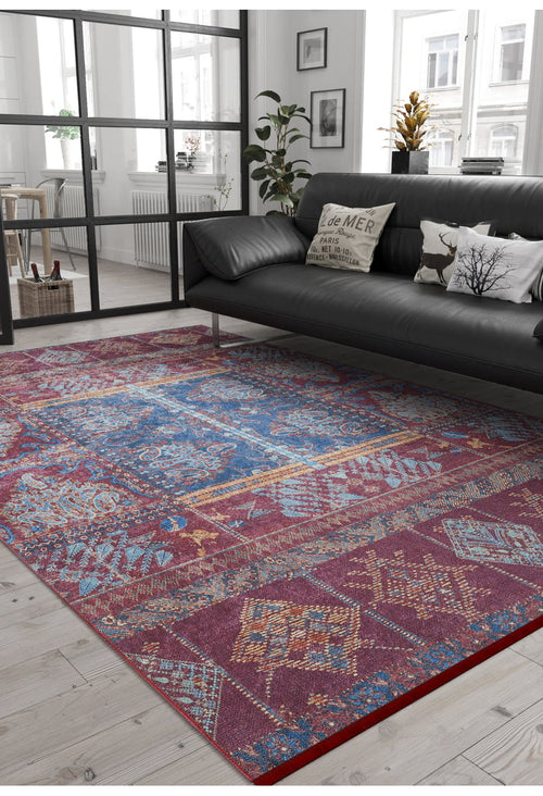 Load image 1 in gallery view. A modern living room with a black sofa, printed pillows, and lush plants features the Majestic Mandarin - Washable Rug - JR1863, ideal for high-traffic areas on a light wooden floor, with large windows letting in natural light.