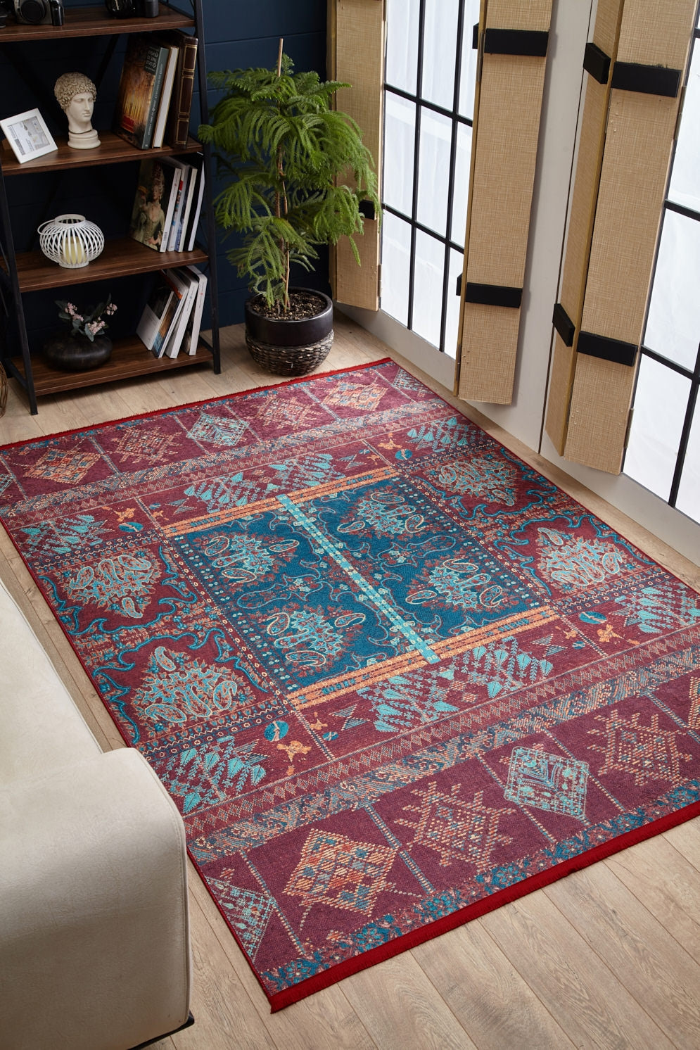 A vibrant living room showcases the Majestic Mandarin - Washable Rug - JR1863 (Custom Size) in red and blue on a wooden floor, with a potted plant, beige sofa, dark bookshelf, and large windows dressed in woven blinds and black-trimmed screens.