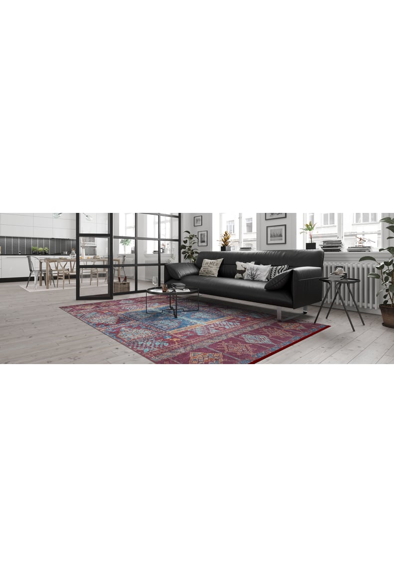 A modern living room with a black sofa, decorative pillows, small round coffee table, and the Majestic Mandarin - Washable Rug - JR1863 (Custom Size) on light wood floors. Large windows brighten the space and the kitchen area is visible in the background.