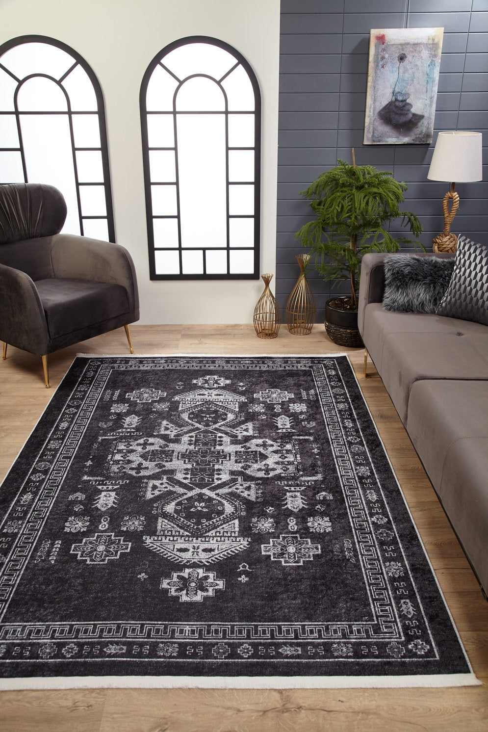 A modern living room features wood flooring, a gray sofa, black armchair, indoor plants, decorative vases, large arched windows with white panes, and the Majestic Symbol - Washable Rug - JR1892 (Custom Size) on the floor beneath a painting.