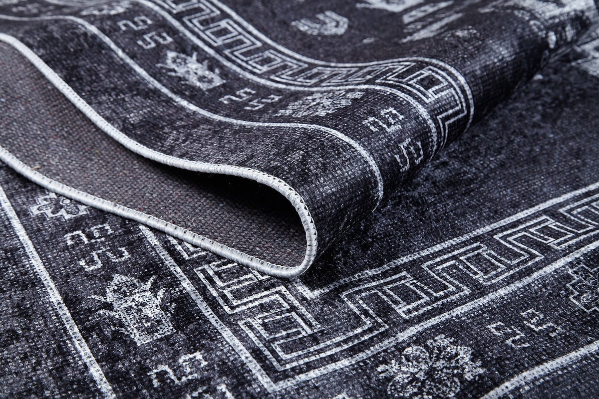 A close-up of the Majestic Symbol - Washable Rug - JR1892 (Custom Size) in black with white geometric and floral patterns, highlighting its folded edge to reveal detailed texture, stitching, and stylish flat weave design.