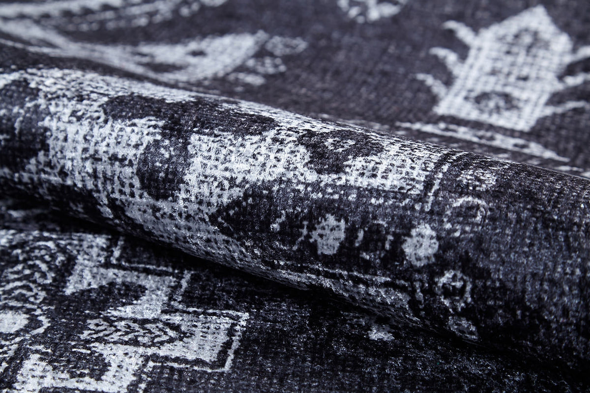 A close-up of the Majestic Symbol - Washable Rug - JR1892 (Custom Size) shows its flat weave fabric in black, gray, and white with a faded geometric pattern. The partially rolled rug highlights its intricate design and textured surface.
