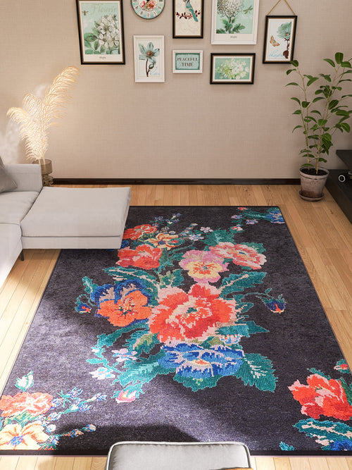 Load image 1 in gallery view. A modern living room with a light gray sectional, wood flooring, framed botanical prints, potted plants in the corners, and the Meadow Dream - Washable Rug - JR2001—a black rug with vibrant red, blue, and green floral patterns ideal for high-traffic areas.