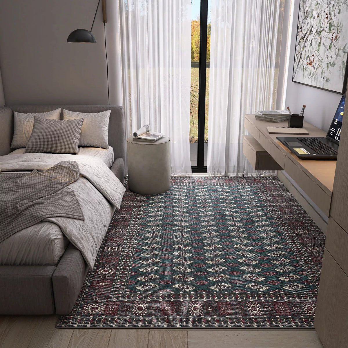 A modern bedroom with a gray bed, pillows, throws, a Midnight Tribal - Washable Rug - JRN1034 perfect for high-traffic areas, a round gray ottoman, desk with laptop, wall lamp, and large windows with sheer white curtains letting in light.
