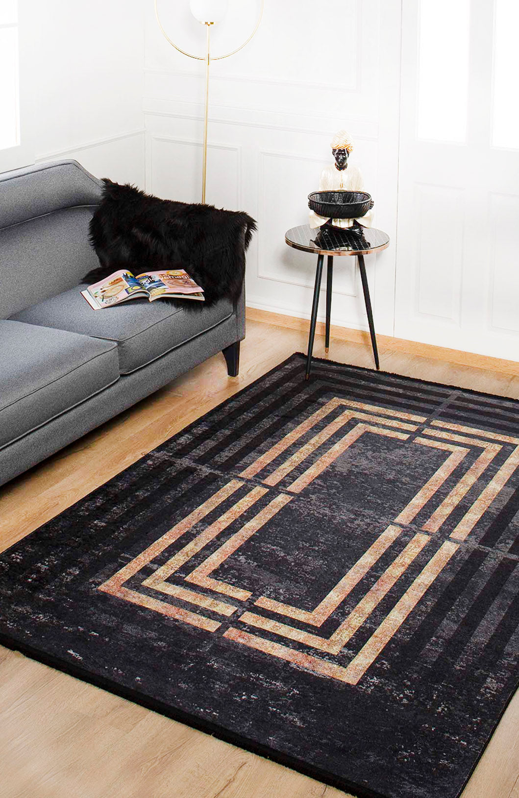 A modern living room with a gray sofa, black faux fur throw, and round side table sits on the Minimalist and Clean - Washable Rug - JR1407 (Custom Size), featuring geometric gold and beige patterns—ideal for high-traffic areas on light wood flooring.