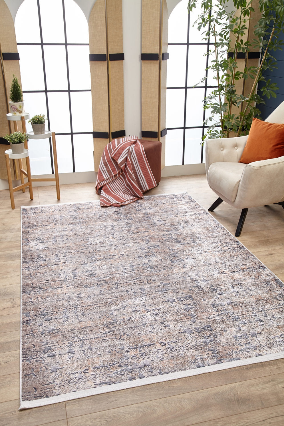 A cozy living room featuring the Modern Enigma - Machine-Washable Rug JR1883 (Custom Size) in cream and beige on a wooden floor, a beige armchair with orange pillows, potted plants, and a striped blanket—perfect for high-traffic areas and easy to clean.
