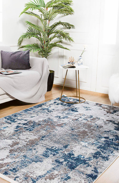 Load image 1 in gallery view. A modern living room featuring the Modern Melody - Washable Rug - JR586 (Custom Size) in blue and gray, a white sofa with gray pillows and blanket, a small round table with a teacup and model ship, a potted palm, and bright window light.