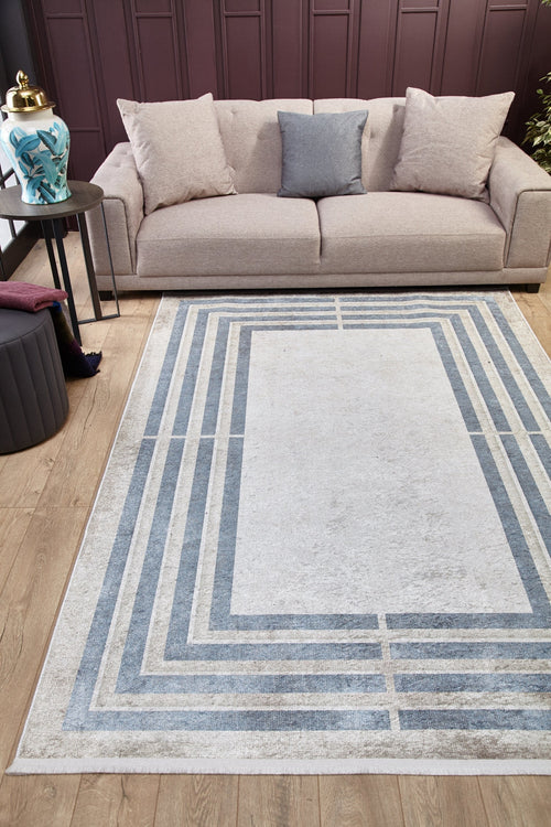 Load image 1 in gallery view. A beige sofa with three cushions sits by a dark paneled wall. In front, the Modern and Regal - Washable Rug - JR1616 (Custom Size) with geometric blue and gray lines lies on a wooden floor, ideal for high-traffic areas, beside a round table with a plant.
