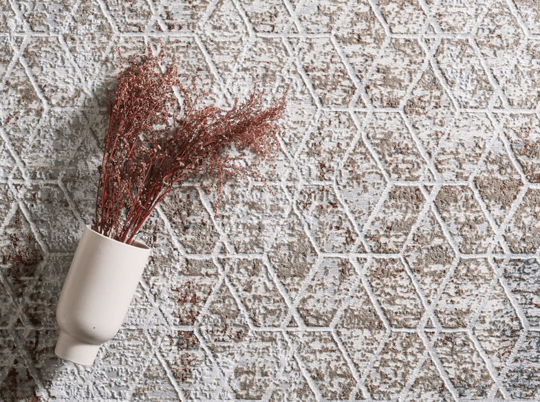 A cream-colored vase holding dried reddish-brown branches lies on its side atop a textured, geometric-patterned surface in white and brown tones.