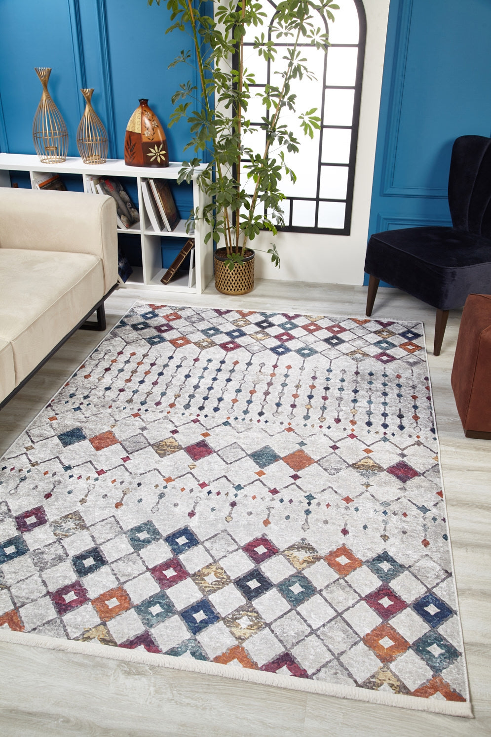 A modern living room with the Moroccan Mosaic - Washable Rug - JR1655 (Custom Size) in gray-anthracite, a beige sofa, dark accent chairs, vases on bookshelves, a potted plant, blue paneled walls, and a tall window—ideal for high-traffic areas.