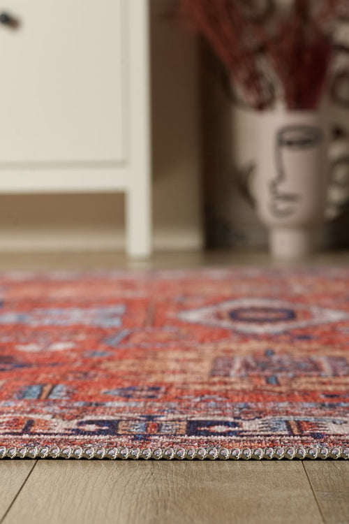 Load image 1 in gallery view. A close-up of the Mystral Kilim-Design Coral Washable Rug - LCC3015 with blue patterns on a wooden floor. A white cabinet and an abstract face vase with dried plants add style—ideal for high-traffic spaces.