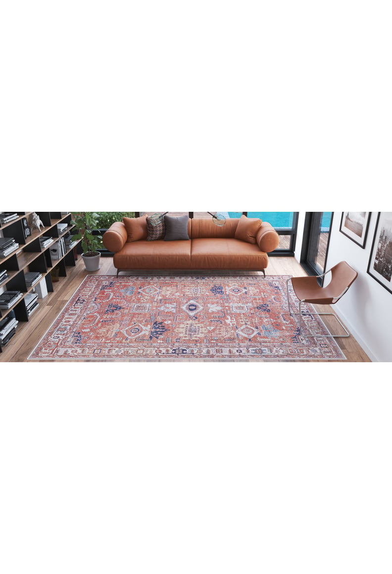 A modern living room with a brown leather sofa, a Mystral Kilim-Design Coral Washable Rug - LCC3015 (Custom Size) ideal for high-traffic areas, a tan minimalist chair, bookshelves, a potted plant, and large windows with natural light.