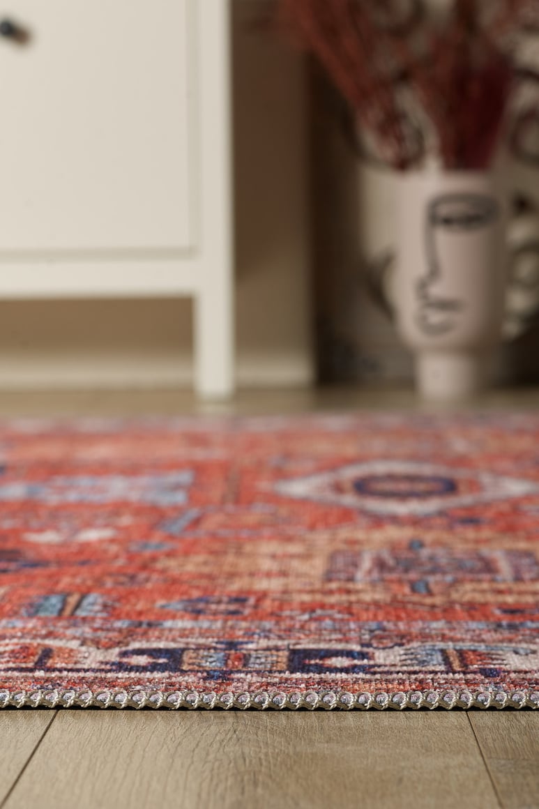 A close-up of the Mystral Kilim-Design Coral Washable Rug - LCC3015 (Custom Size) on a wooden floor, ideal for high-traffic spaces, with a blurred white cabinet and abstract face vase in the background.