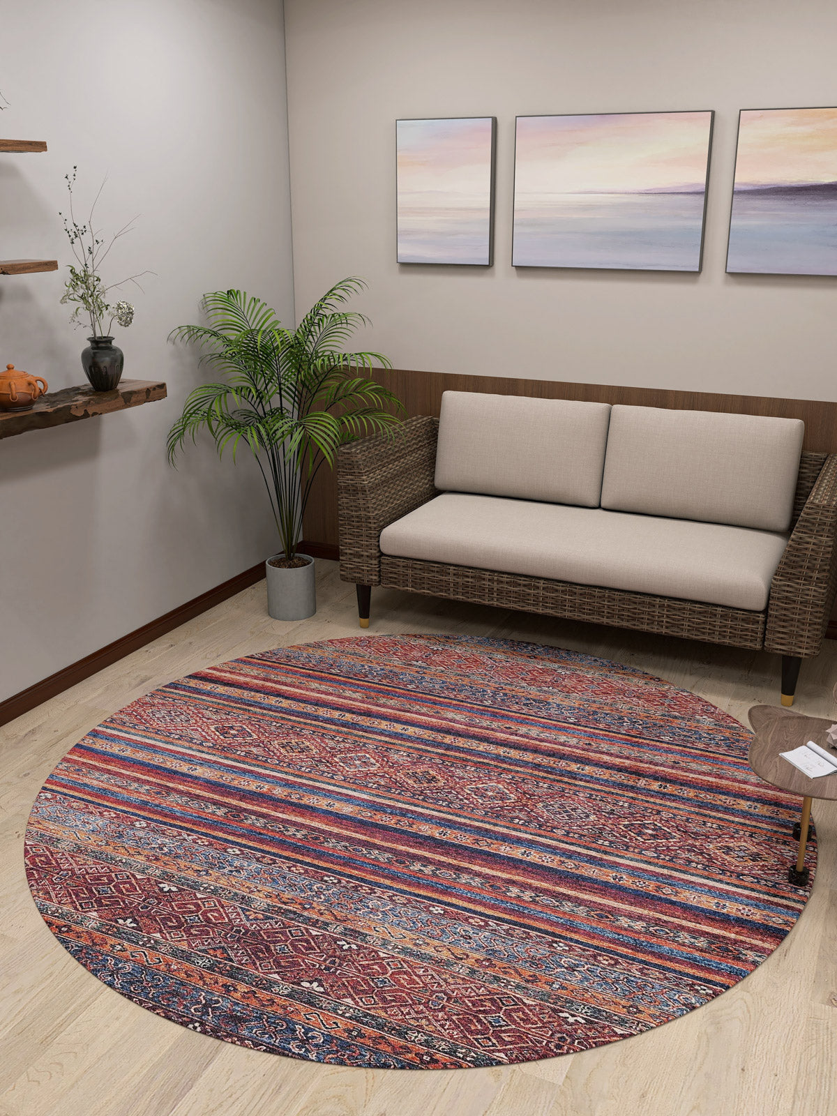 A cozy living room with a beige loveseat, a Native Nightscape - Washable Rug - JR5093 (Custom Size) featuring red and blue stripes, a potted plant, pastel landscape paintings, and wooden shelves holding small decor.