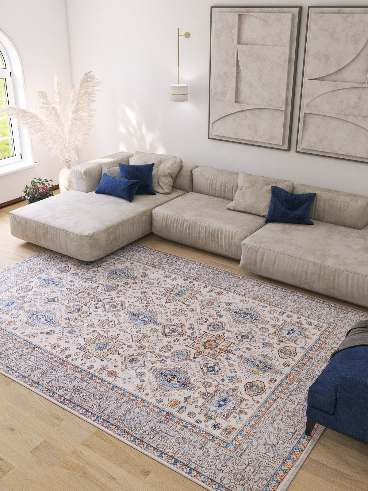 A spacious living room featuring a large beige sectional, blue accent pillows, the Nexa Bordered Beige Washable Rug (LCC3009), light wood floors, tall pampas grass in a vase, and abstract wall art above the couch.